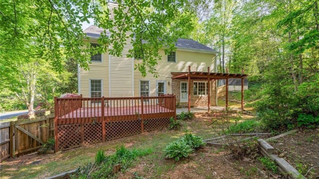 A redwood deck with a pergola, yellow house, and lush forest backdrop with a wooden fence.
