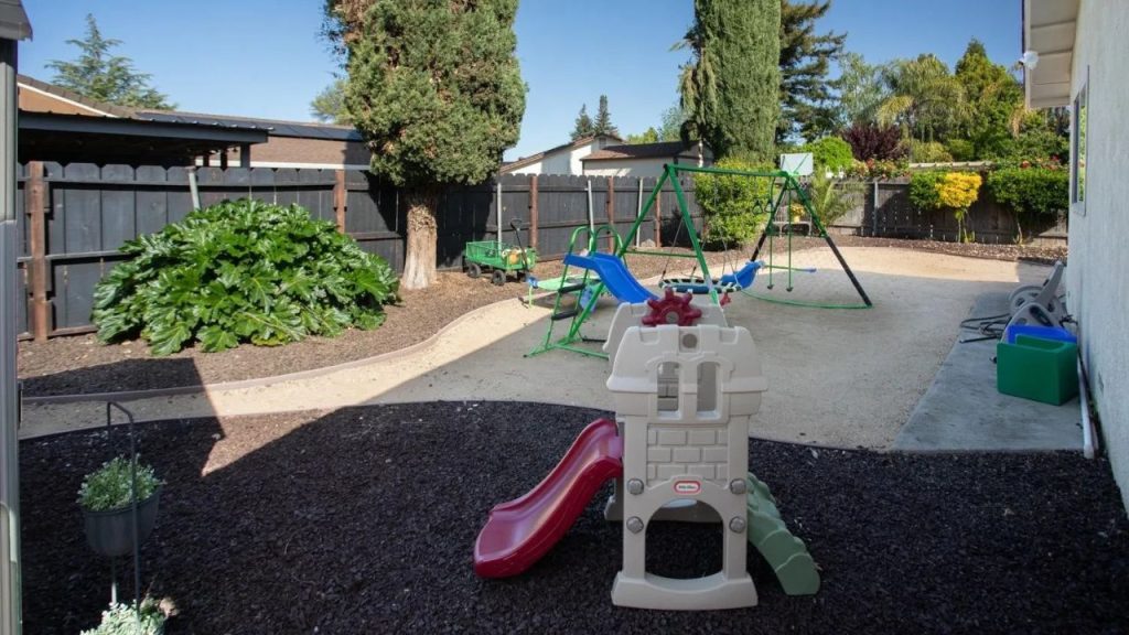 A sunny backyard play area with a playground set, red slide, castle structure, and surrounding greenery.