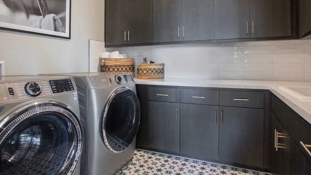 A modern laundry room with dark cabinets, white countertop, subway tile backsplash, a washer and dryer, woven baskets, patterned black-and-white floor tiles, and a framed photo on the wall.
