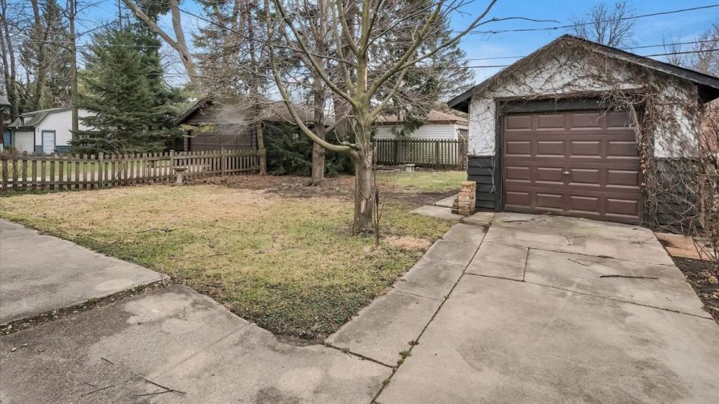 A quaint backyard with a grassy lawn, wooden fence, bare trees, a driveway, and a rustic garage with dark wood and climbing vines under a clear blue sky.