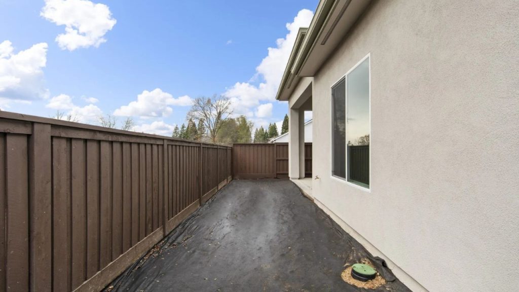 A backyard with a wooden fence, a modern home exterior with large windows, and an open sky with scattered clouds.
