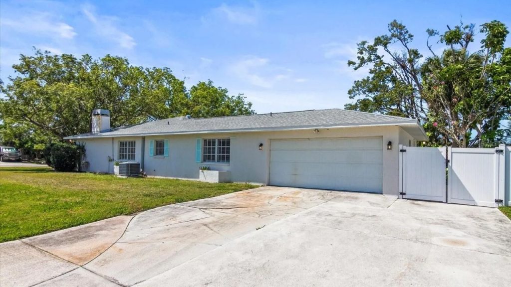 A single-story suburban home with a light gray exterior, shingle roof, white garage door, pale blue shutters, and a white fence surrounded by lush trees.
