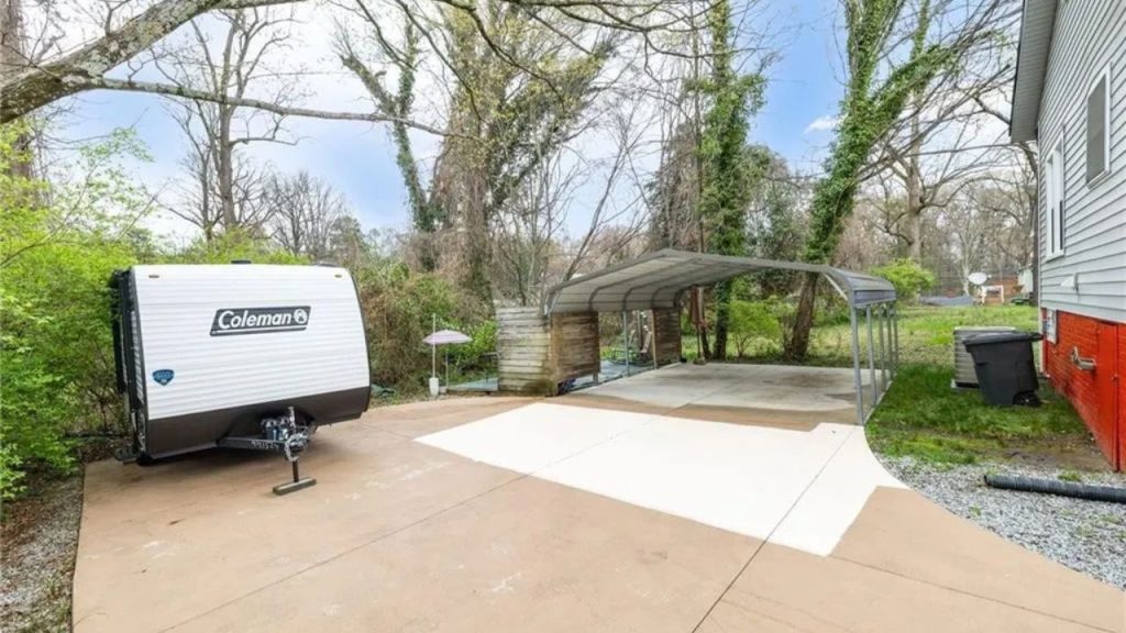 Backyard driveway with carport and a Coleman camper in a wooded setting.