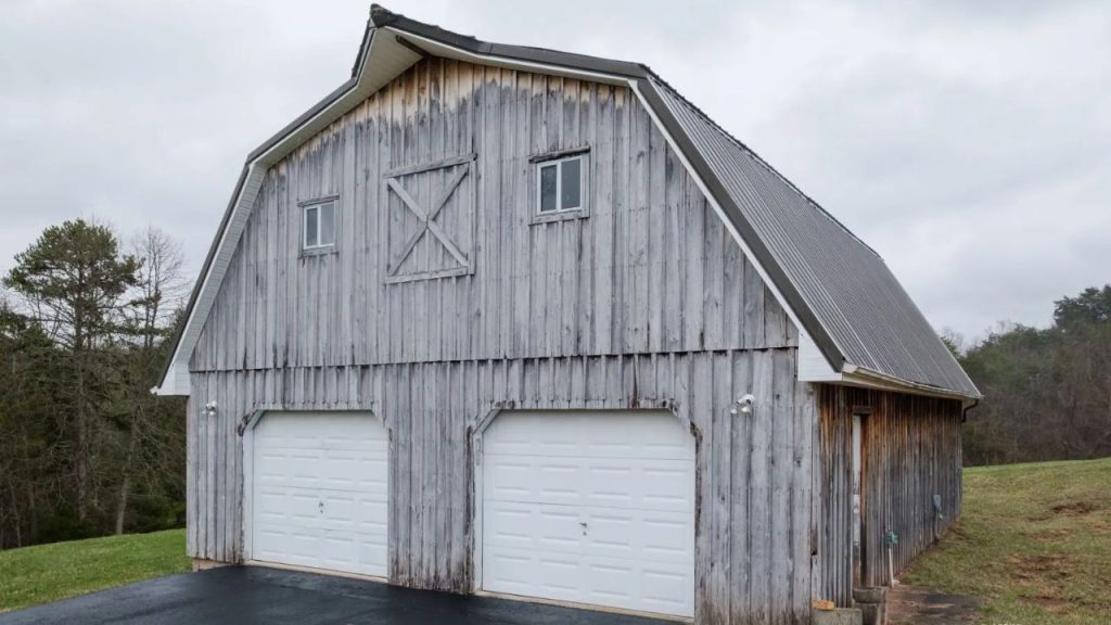A weathered wooden barn-style garage with a metal roof, white doors, and a blacktop driveway in a rural setting.