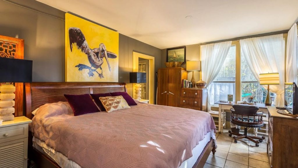 A cozy bedroom with a wooden bed, pink quilt, purple pillows, a bold pelican painting, and a desk by large windows with sheer curtains.