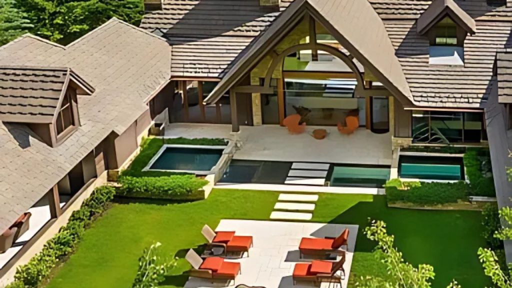 A rustic patio with orange lounge chairs, an infinity pool, and a second pool, surrounded by green lawns and a wooden-accented house.