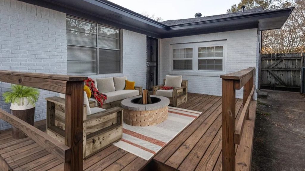 A cozy deck with wooden furniture, a stone fire pit, and a striped rug, framed by house windows.