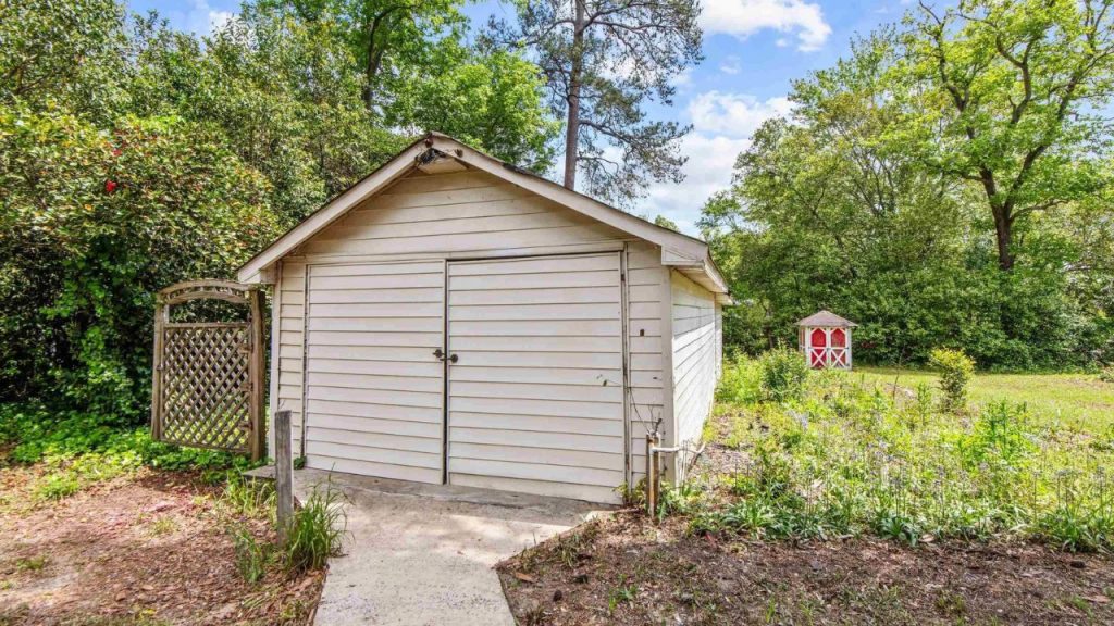 Beige garden shed with double doors and a red playhouse in a wooded backyard.