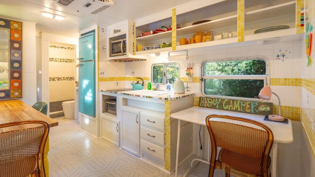 A retro camper kitchen with a turquoise fridge, yellow-trimmed cabinets, wicker chairs, and a cozy dining nook by a window.