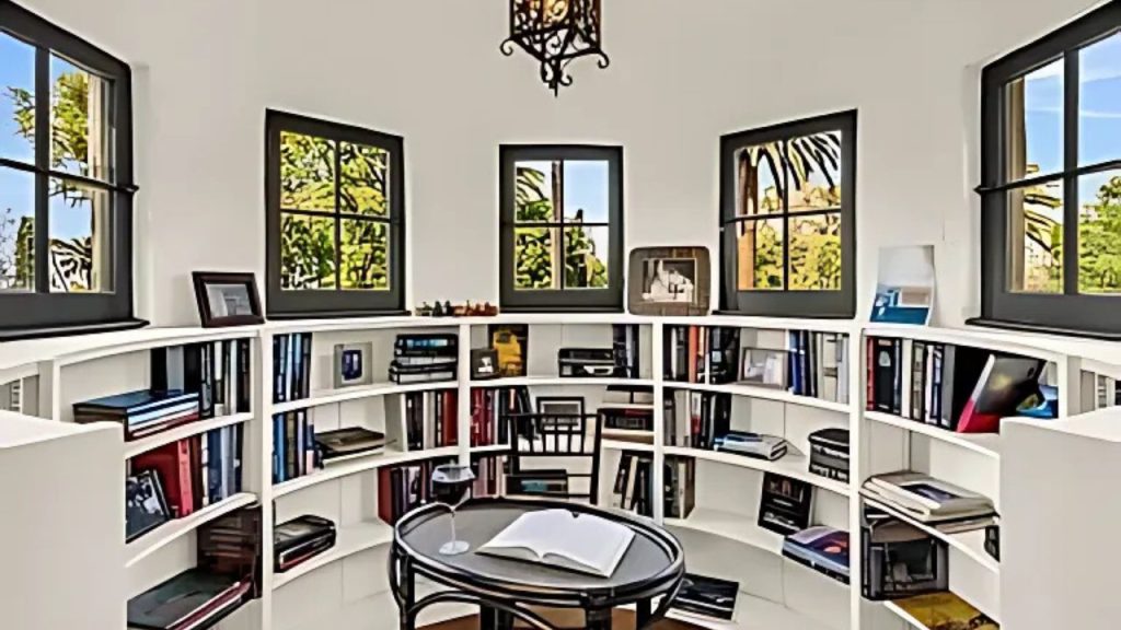 A cozy reading nook with curved bookshelves, a round table, and large windows overlooking greenery.