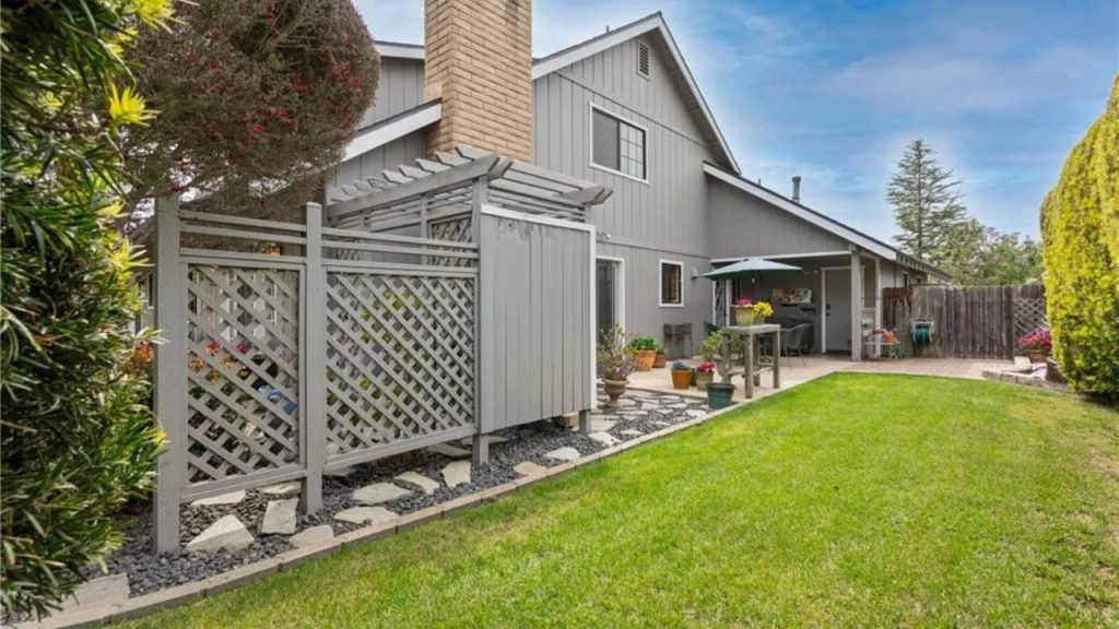 A tranquil backyard with green grass, a pergola, patio with plants, and a gray house.