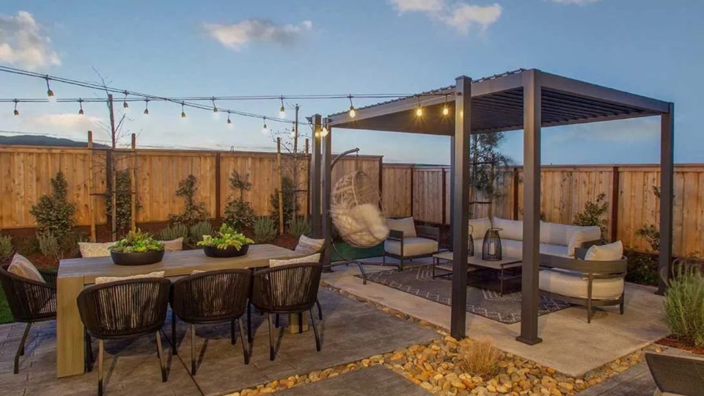 A backyard patio with a pergola, string lights, a wooden dining table with wicker chairs, a lounge area with a sectional and hanging egg chair, wooden fencing, and potted plants.