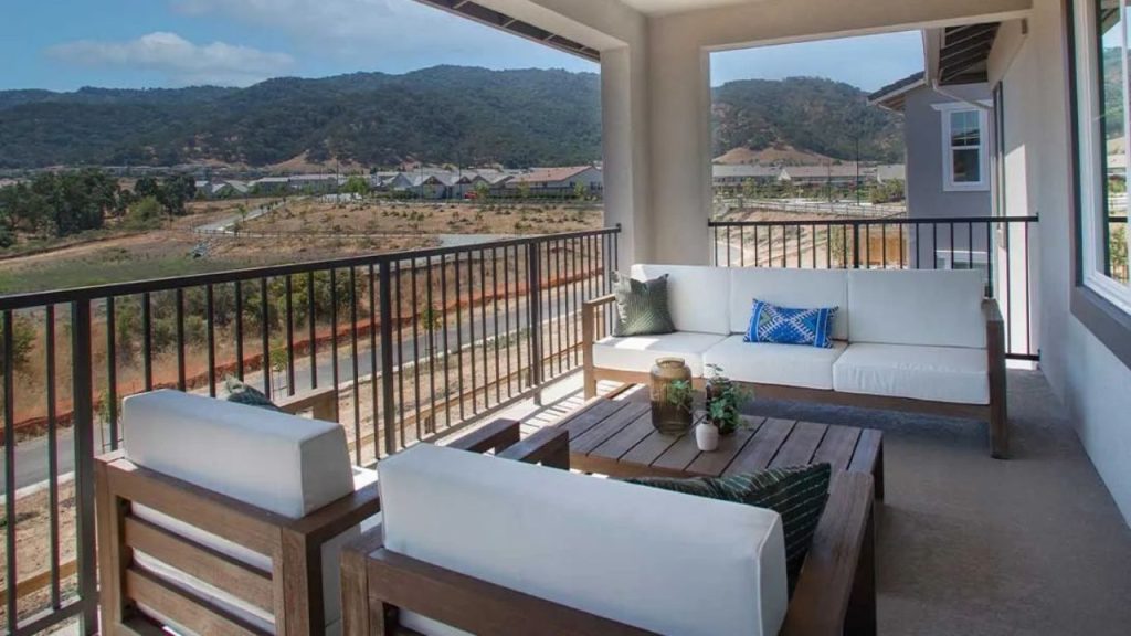 A covered patio with a white sofa, wooden chairs, a coffee table, and a view of rolling hills and distant mountains.