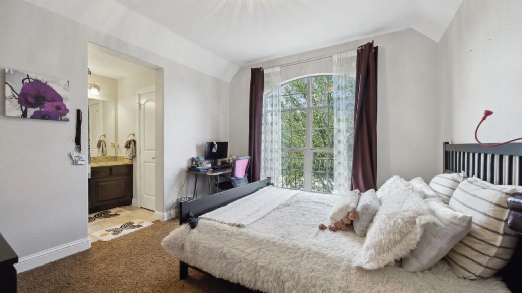 Sunny bedroom with arched window, plush bed, and cozy desk setup.