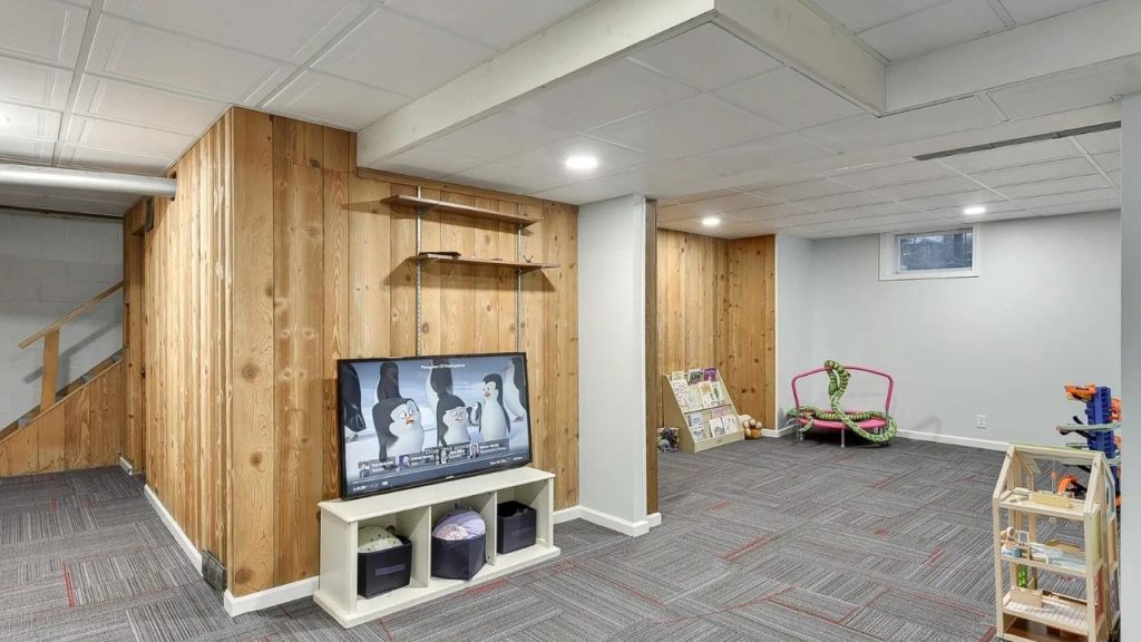 Basement playroom with wood paneling, TV, toys, and soft carpet tiles.