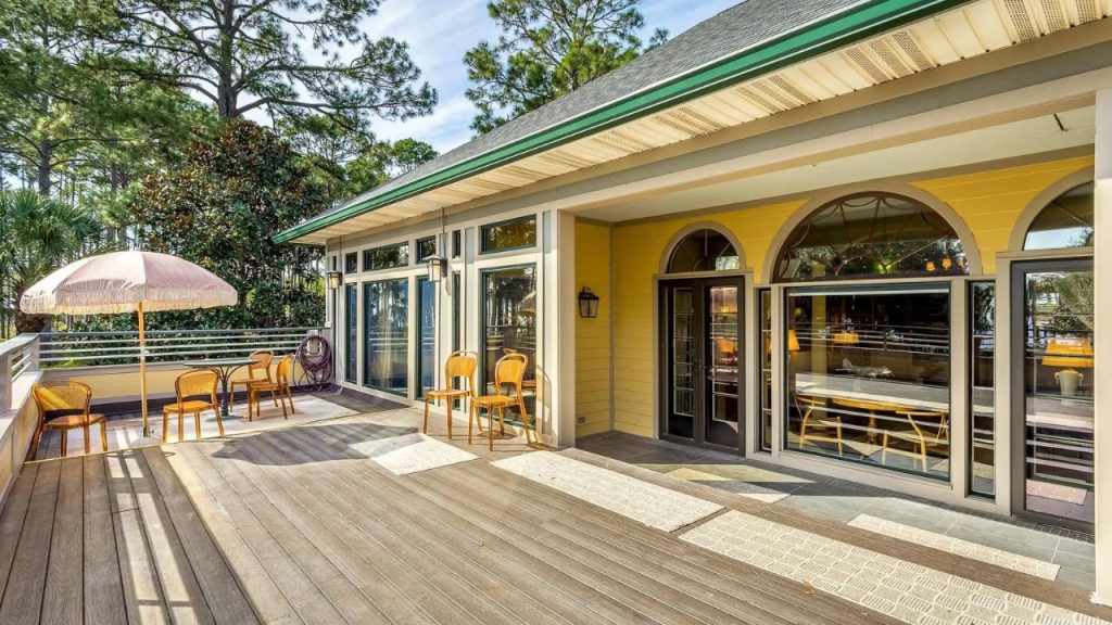 A sunny deck with wicker chairs, a pink umbrella, and a yellow building with arched windows surrounded by trees.