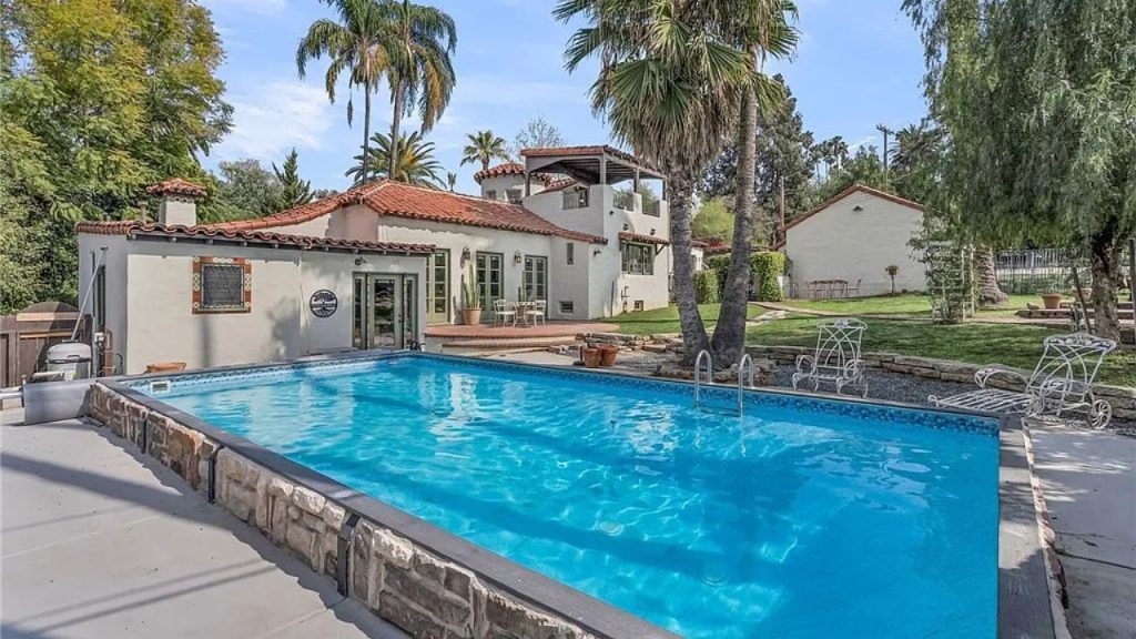 A serene poolside with a Spanish Colonial home, palm trees, and a cozy patio under a bright sky.