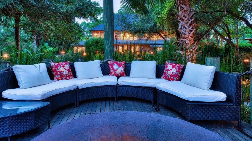 A tropical patio with a wicker sofa, red floral pillows, a fire pit, and a house surrounded by lush greenery.