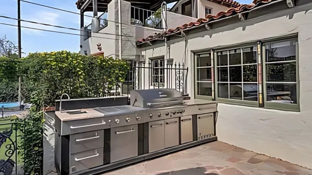 A stylish outdoor kitchen with a stainless-steel grill, surrounded by greenery and a Spanish Colonial home.