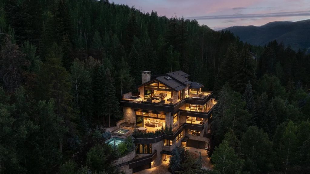 A luxurious home nestled in a forest, glowing at twilight with a hot tub and mountain views.