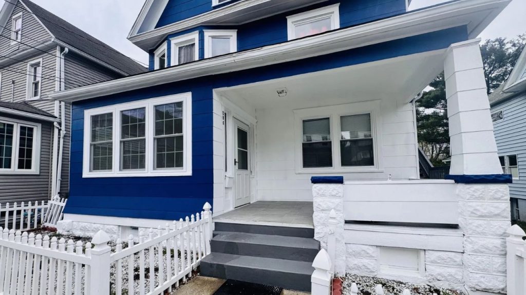 A charming blue house with a white porch and picket fence, featuring large windows and stone steps under a cloudy sky.