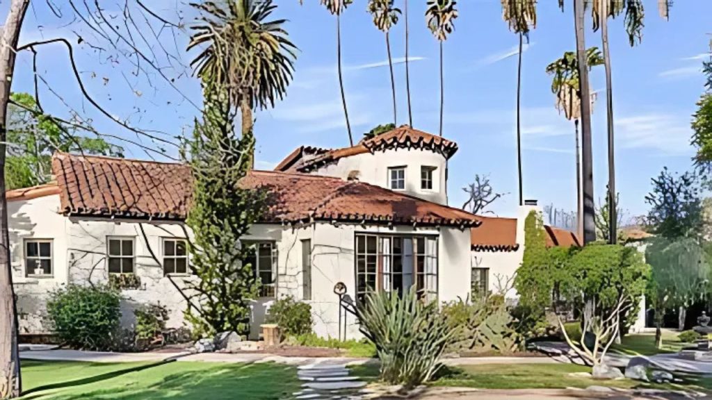 A charming Spanish Colonial home with a red-tiled roof, white stucco walls, and a circular tower, surrounded by palm trees and a lush lawn.