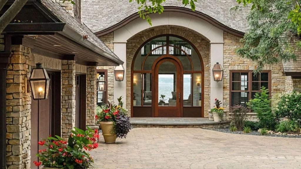 A charming stone entrance with an arched doorway, flanked by lanterns and vibrant flower pots, opening to a lake view.