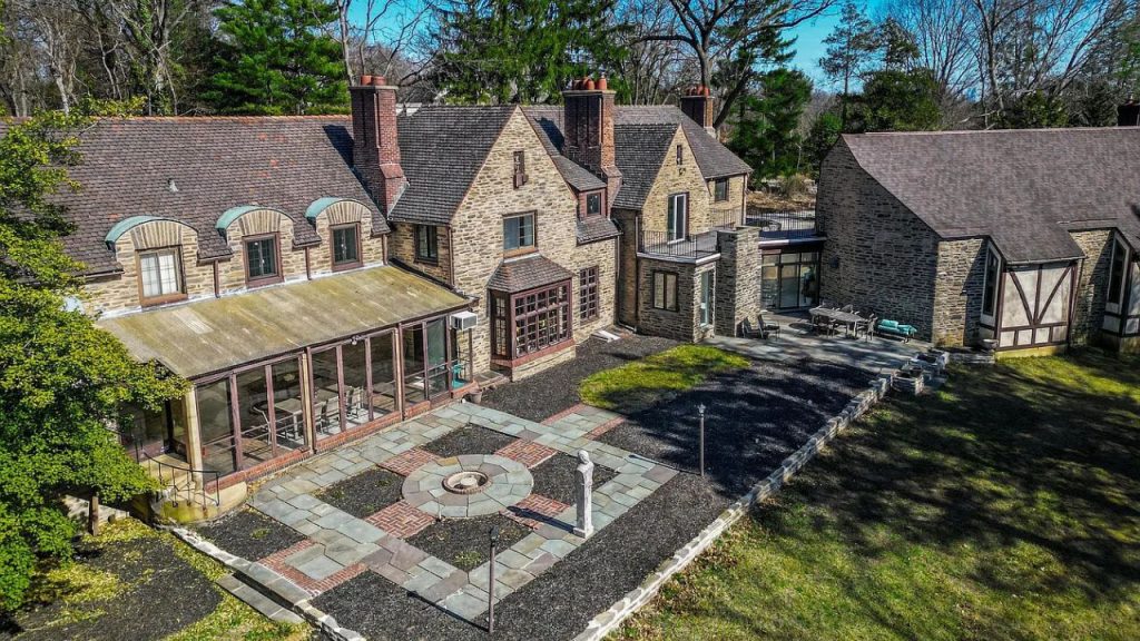 A stunning stone patio with a fire pit, surrounded by a historic stone house and lush trees under a clear sky.