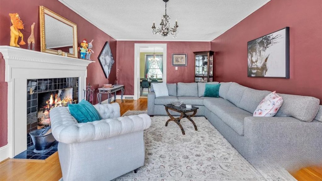 A cozy living room with a fireplace, gray sectional sofa, teal pillows, and a chandelier, featuring burgundy walls and eclectic decor.