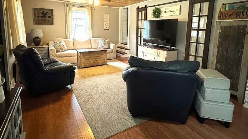 A cozy living room with leather chairs, a beige sofa, wooden floors, and a TV, lit by natural light from a large window.