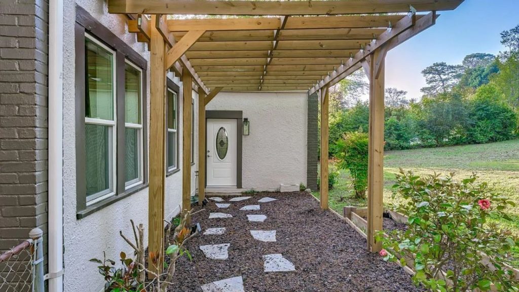 A charming covered entry gate with wooden beams, stone path, and lush greenery leading to a white door.