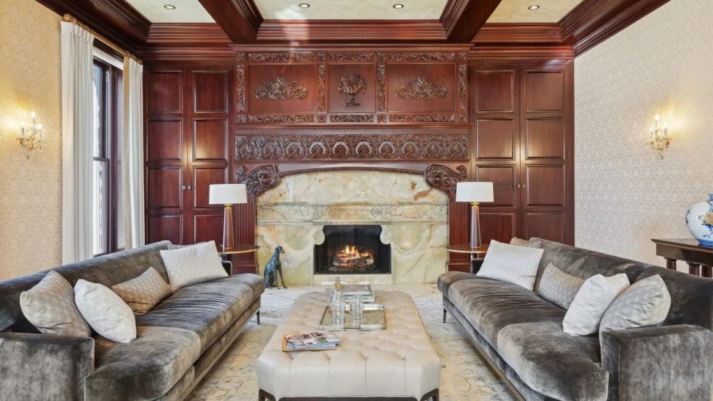 A luxurious living room with gray sofas, a marble fireplace with intricate mahogany paneling, and a tufted ottoman, lit by soft wall sconces and lamps.