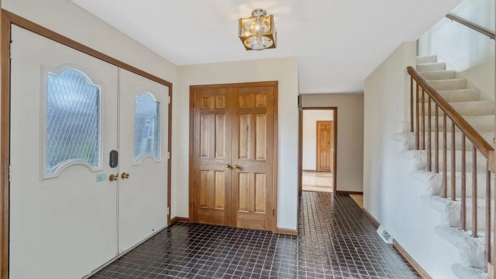 Bright entryway with black tile flooring, wooden double doors, and a staircase with carpeted steps.