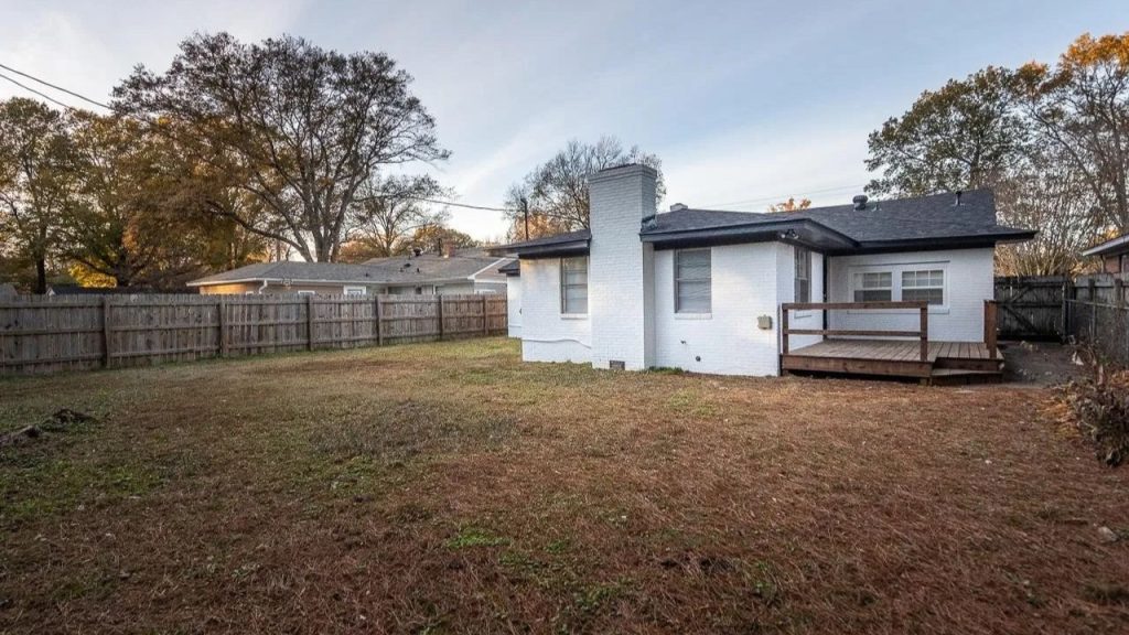 Spacious fenced backyard with a small wooden deck and white brick exterior.