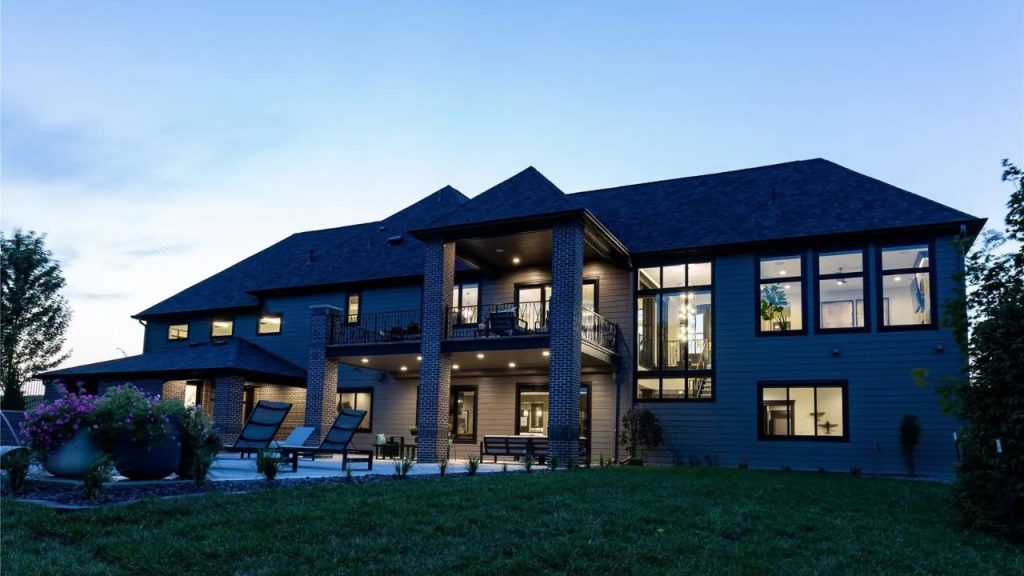 A modern home’s back exterior at dusk, featuring large windows, a balcony, covered patio with seating, and a well-kept lawn with potted plants.