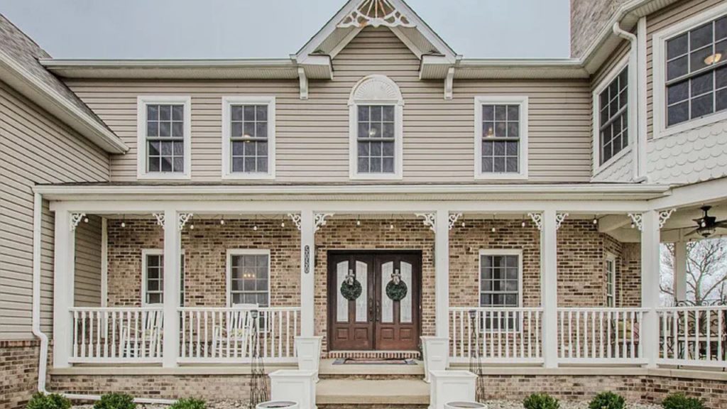 A welcoming front porch with white columns, string lights, and double doors, framed by brick and beige siding.