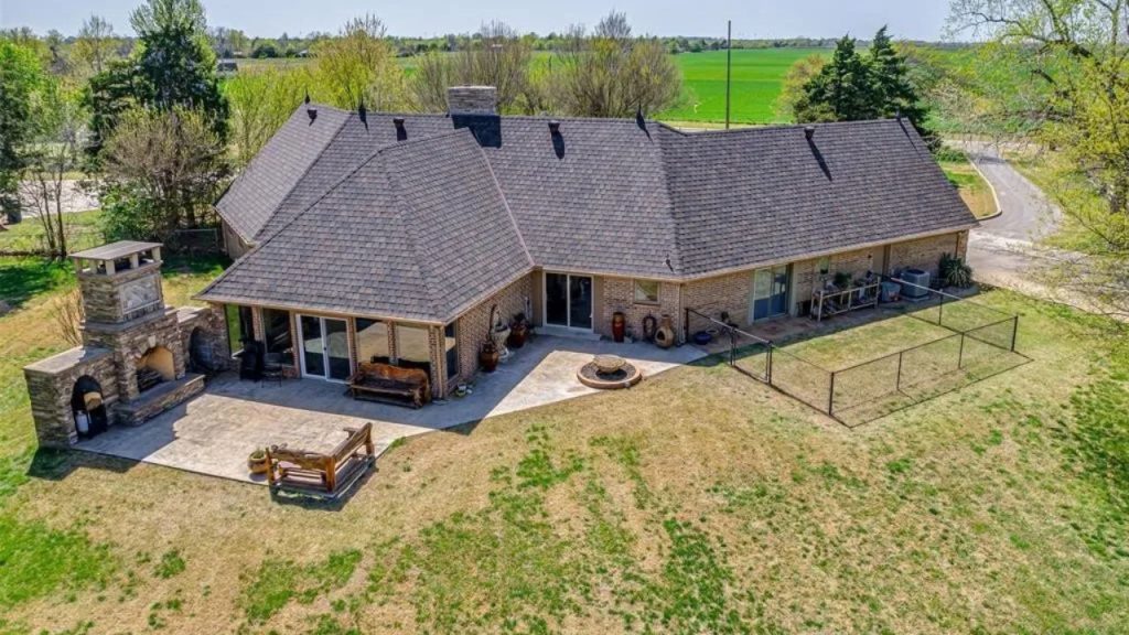 Aerial view of a colonial-style home’s backyard with a stone fireplace, fire pit, and fenced area, surrounded by lush countryside.