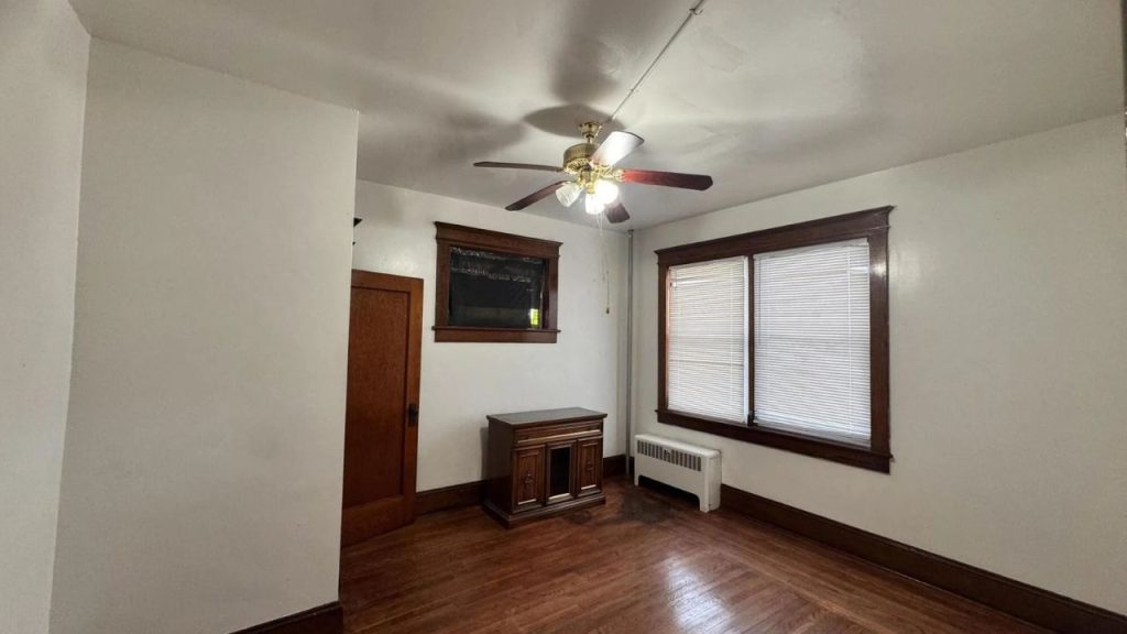 A bright living room with wooden floors, a ceiling fan, large windows, and a wooden cabinet.