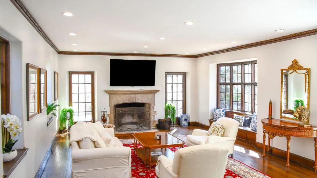 A cozy living room with a stone fireplace, white sofas, and large windows, accented by a red rug and elegant decor.