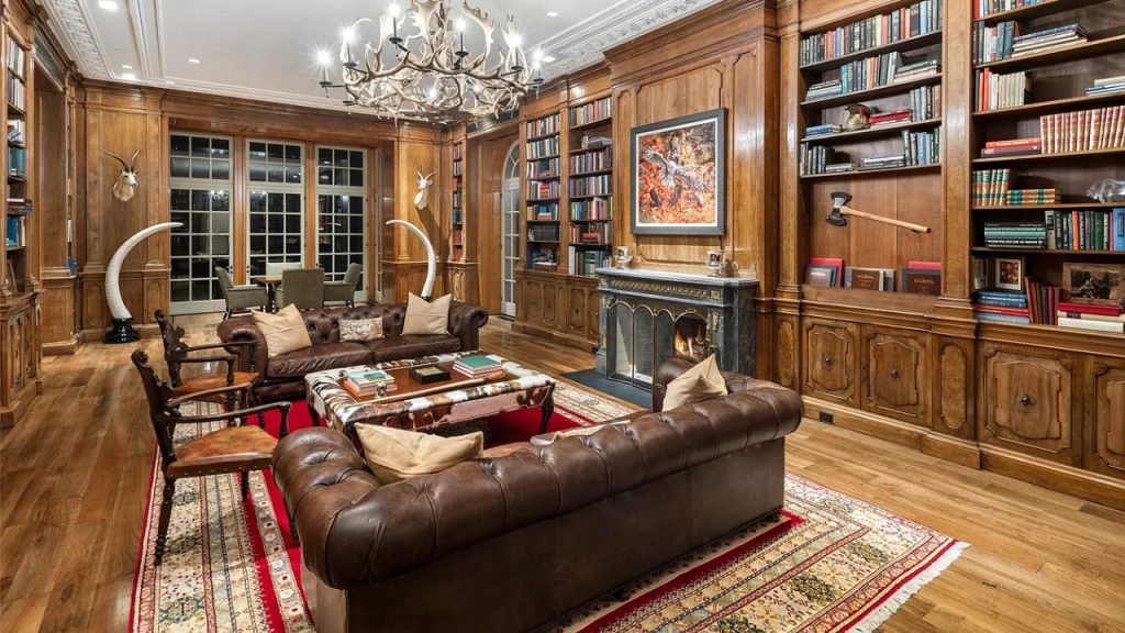A cozy library living room with wooden bookshelves, leather sofas, a fireplace, and a chandelier.