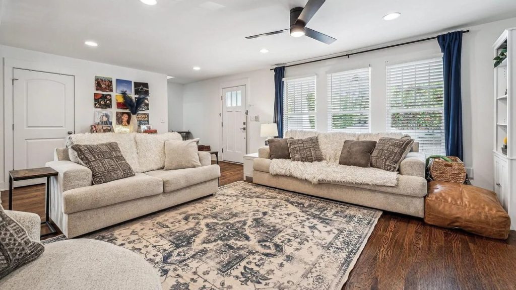 A cozy living room with beige sofas, a patterned rug, and large windows with blue curtains.