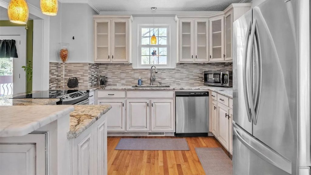 A bright kitchen with white cabinets, marble counters, stainless steel appliances, and a mosaic tile backsplash, accented by pendant lamps and a cozy rug.