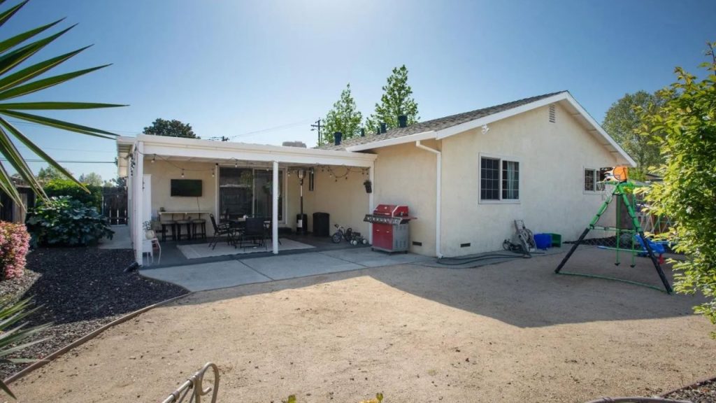 A sunny backyard patio with a covered seating area, barbecue grill, swing set, and lush greenery, attached to a light-colored house.