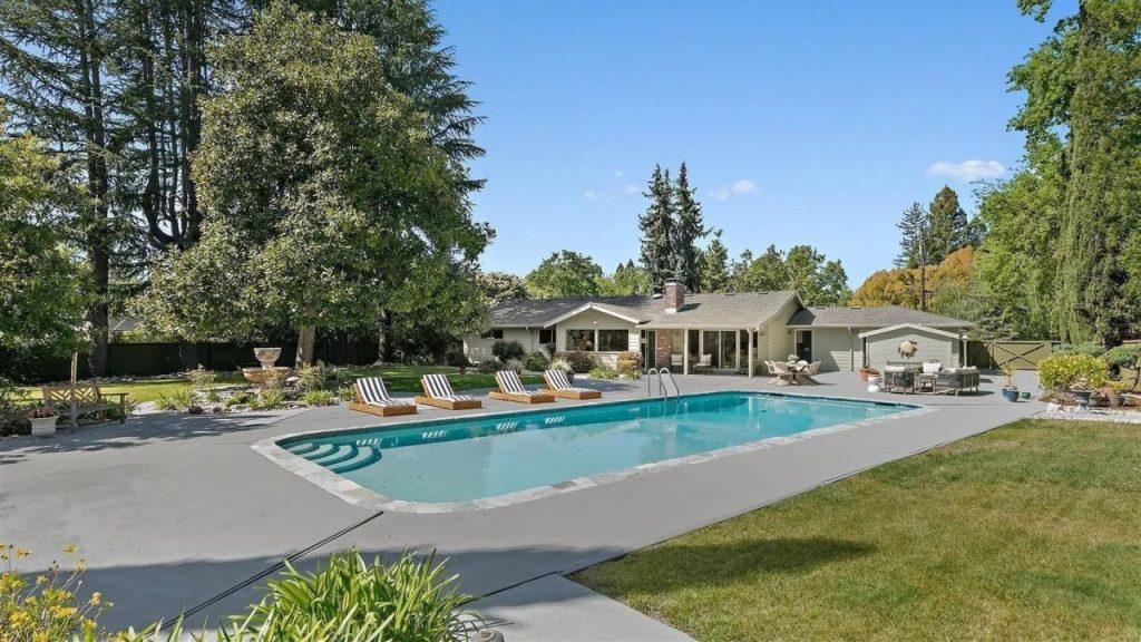 A serene backyard with a sparkling pool, lounge chairs, and a cozy patio, framed by tall trees and a stylish home.