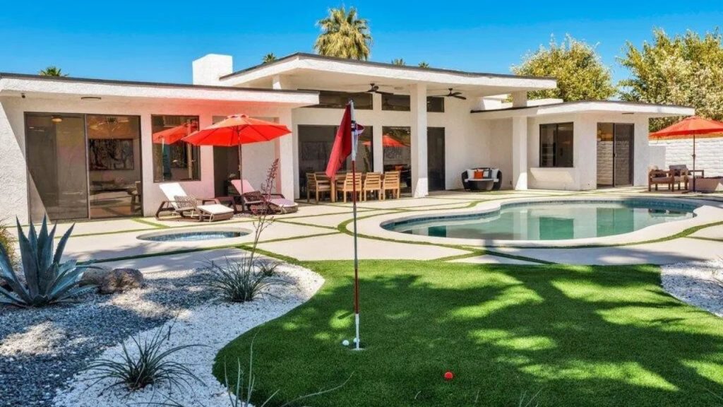 A stylish mid-century modern backyard with a pool, hot tub, putting green, and red umbrellas, nestled among desert plants.