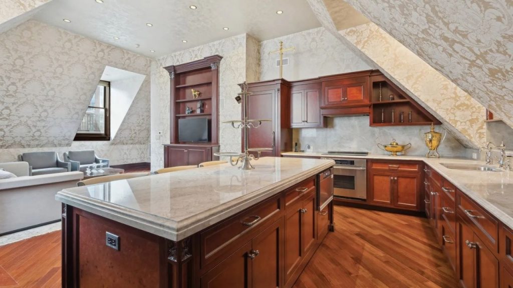 An elegant kitchen with mahogany cabinets, marble countertops, and a large island, featuring a cozy seating area under an angled ceiling with patterned wallpaper.