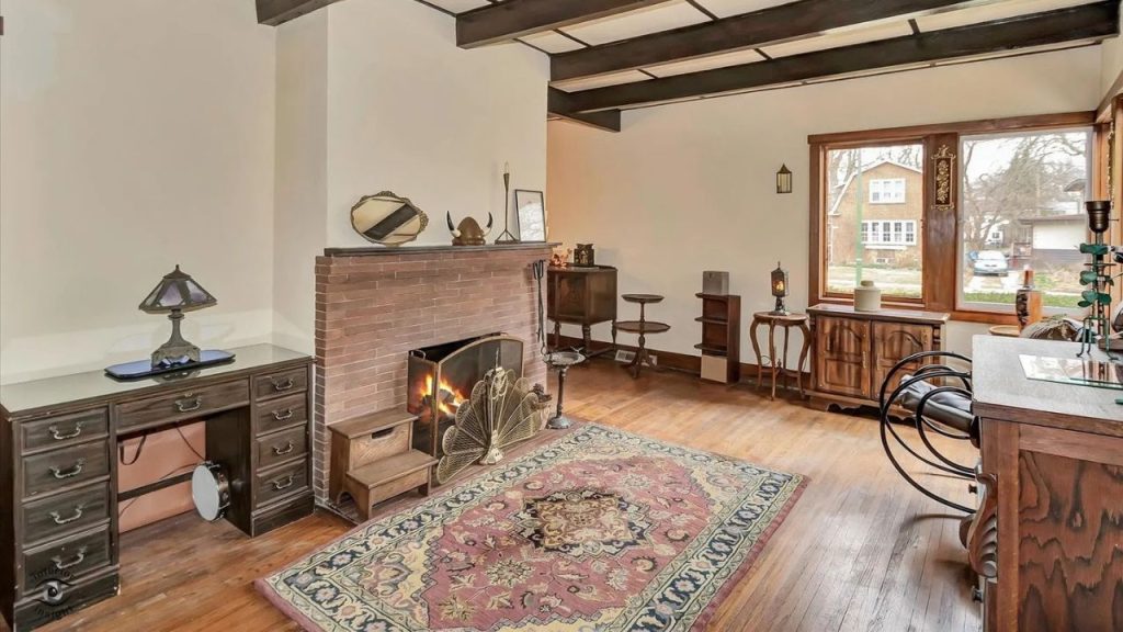 A cozy living room with a brick fireplace, Persian rug, wooden beams, antique desk with a Tiffany lamp, and large windows overlooking a quiet neighborhood.
