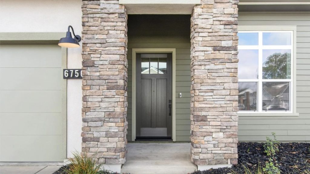 A modern home entryway with stone columns, a gray door with frosted glass, a large window, and a lantern light, accented by grasses in black mulch.
