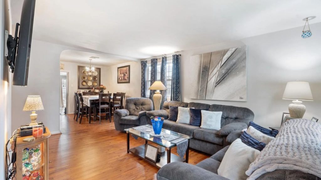 Cozy living room with gray sofas, glass coffee table, lamps, and a dining area in the background.