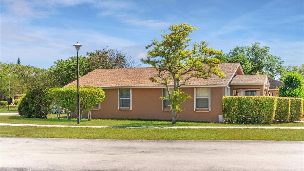 A peaceful backyard with a tan house, green hedges, a tall tree, and a streetlamp under a clear blue sky.